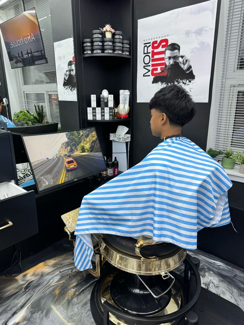 Client in a striped barber cape playing a racing game at Mori Cuts
